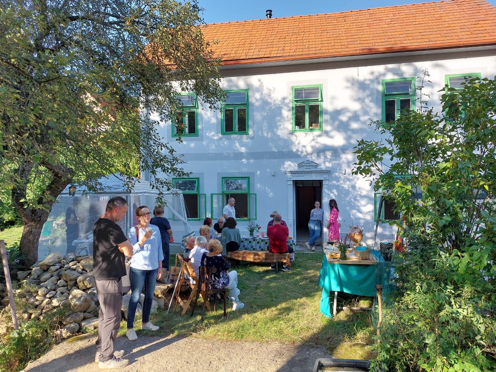Návštevníci sedia a rozprávajú sa v záhrade pred bielou budovou Galérie Ambroze so zelenými oknami a červenou strechou